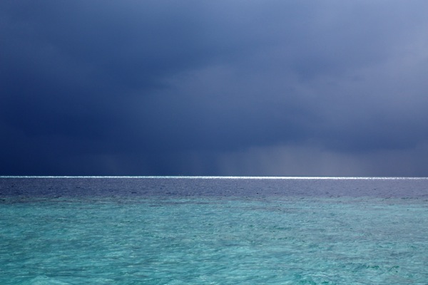A photograph showcasing distinct horizontal layers: a textured expanse of turquoise water at the bottom, topped by a thin, bright horizon line, above which stretches a vast, dark blue-grey sky filled with heavy clouds. The composition emphasizes these stratified layers, mirroring the theme of the post suggested by phrases like "layer cake" and the concept of "lateral" perspectives, where different levels or aspects are considered side by side. The image evokes a sense of depth and the interplay between different atmospheric and aquatic strata. This photo was taken in the Maldives.