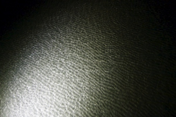 A close-up, high-contrast, black-and-white image of a patch of sand, illuminated by a single, focused light source. The texture of the sand is sharply defined, creating a pattern of small ridges and depressions that strongly resembles a human fingerprint or thumbprint, alluding to the post's title and its exploration of the human 'print' or mark on nature. The light casts a shadow that emphasizes the intricate, repeating patterns.