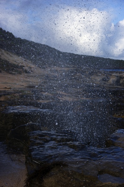 A close-up, high-speed photograph of fine ocean spray and water droplets rising from a blowhole or breaking wave. The image's title, 'spittle,' directly connects to the visual of the water spray and continues the running theme of the 'Coastal Tranche' series, which explores the various forms of spray and foam from the Cape to Cape coastline.