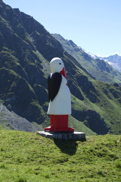 A photograph of the sculpture titled "Baby Bringer" by artist Donna Dodson, standing on a grassy hill in the Verbier 3D Sculpture Park in the Swiss Alps. The sculpture is a figurative representation of a white, stork-headed goddess of motherhood, pregnant and celebrating fertility. The piece stands upright on a perch overlooking the valley, with a steep, green mountain range visible in the background under a cloudy sky.