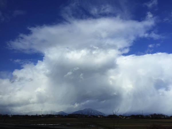 A dramatic sky filled with swirling, textured clouds under a bright blue expanse. The cloud formation appears to be a localized storm or weather event, contrasting with the typically clear or softly clouded skies often associated with the region. Below the intense sky, a flat landscape with some distant trees and possibly buildings is visible along the horizon line, emphasizing the powerful and unusual cloud formation above.
