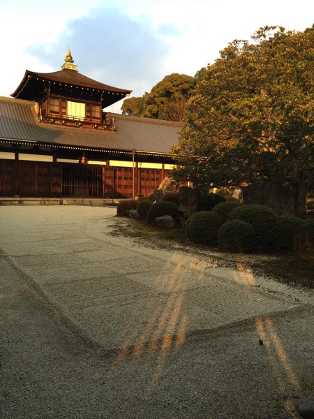 A photograph of a traditional Japanese temple building with a tiered roof, captured in the warm light of late afternoon. A strong, striped pattern of sunlight is cast on the paved path in the foreground, creating a striking "lightbox" effect. 