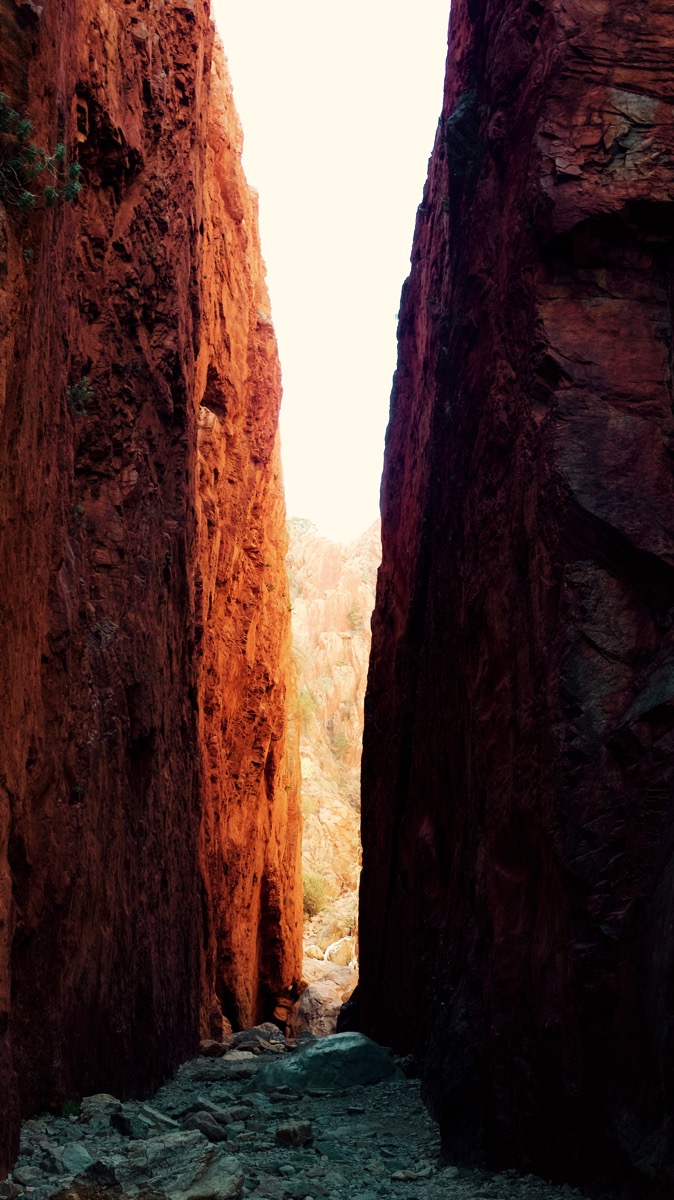 A vertical photograph looking up from the floor of Standley Chasm, also known as Angkerle Atwatye, in the Northern Territory of Australia. Two towering, reddish-orange rock walls rise steeply on either side, their vertical textures emphasized by the narrow strip of bright daylight that enters from above. The light powerfully illuminates the sandy ground at the base of the chasm and the top of the rock faces, while the rest of the walls are cast in deep shadow. The image conveys a dramatic sense of scale and enclosure within this significant geological feature of Central Australia.