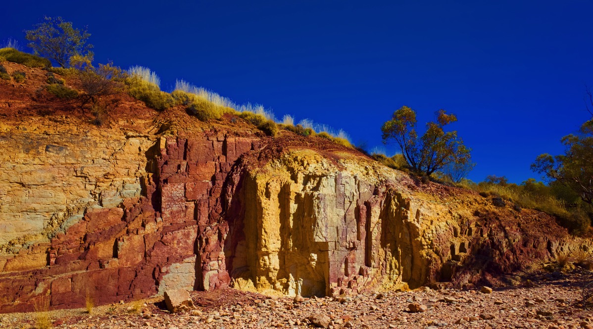 A photograph of the Ochre Pits in Australia, featuring a detailed view of a layered cliff face. The rock is a tapestry of vibrant, naturally occurring ochre, with prominent bands of yellow, deep orange, and earthy red. Vertical streaks of color run down the face, suggesting the 'leaching' process mentioned in the title. The top of the formation is capped with a few small trees and dry grasses, all set against a clear, brilliant blue sky. The afternoon light sharply defines the rich textures and colors of the mineral deposits.
