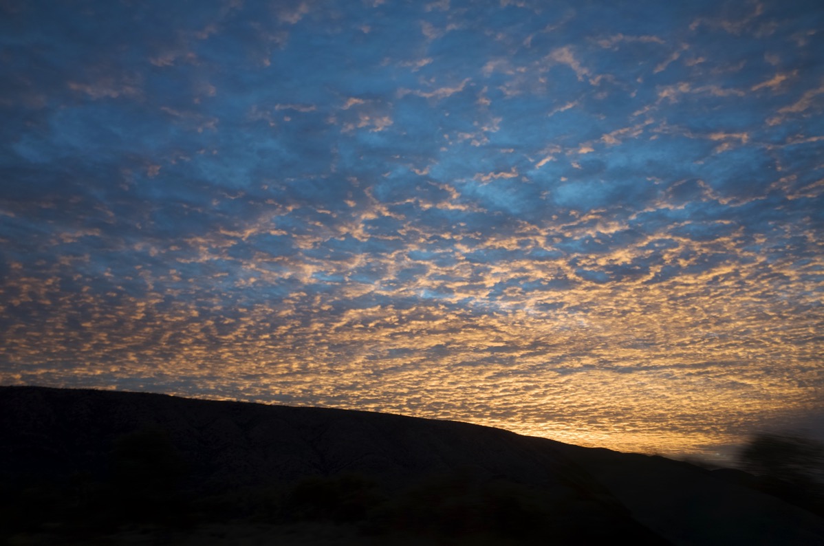 A wide-angle landscape photograph of the Larapinta Range against a dramatic and cloudy sunrise sky. The sky is filled with streaks of gold and blue, with ominous clouds gathering overhead. The image was taken from Namatjira Drive. 