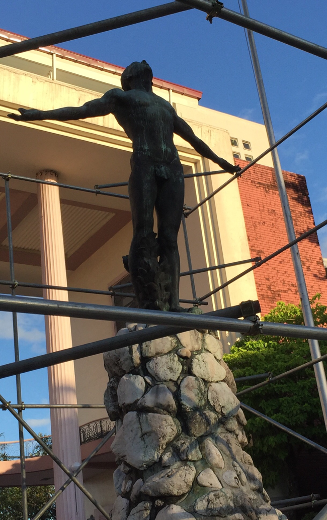 A photograph of the Rizal Monument in Manila's Rizal Park, a memorial dedicated to the Philippine national hero, Dr. José Rizal. The monument features a bronze statue of Rizal on a granite pedestal. In this photograph, the statue is surrounded by scaffolding, likely for the cleaning of the statue.