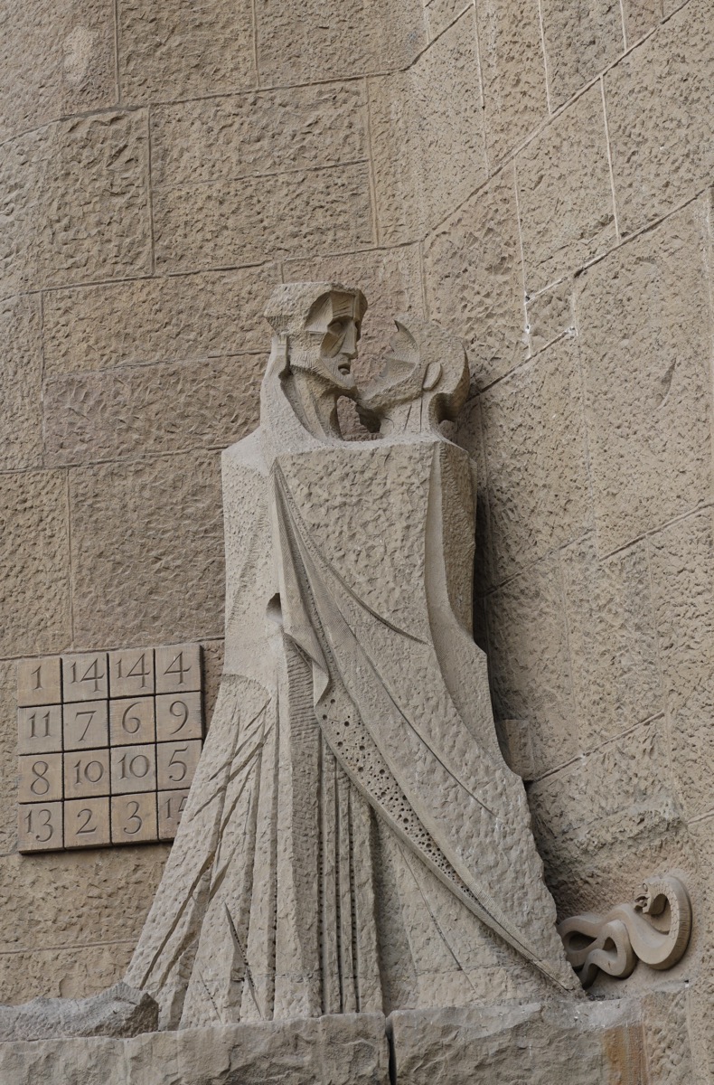 A photograph of the stone sculpture 'The Kiss of Judas' by Josep Maria Subirachs, on the Passion Façade of the Sagrada Família in Barcelona, Spain. The sculpture depicts Jesus and Judas in a moment of betrayal, with a famous magic square in the background whose numbers always add up to 33.