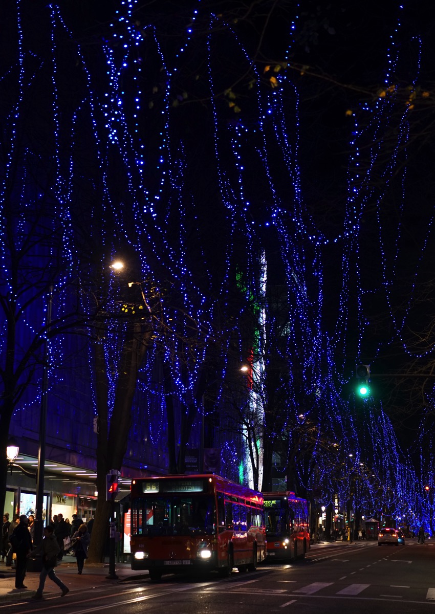 A nighttime street scene in Bilbao, Spain, bathed in the rich, deep blue light from strings of lights draped between trees. The vibrant blue illumination, which is reminiscent of fountain pen ink, contrasts with the warm interior lights of the buildings and the passing red bus. The image captures the magical atmosphere of the city at night, reflecting the poem's wordplay on 'quink' and 'ink'.