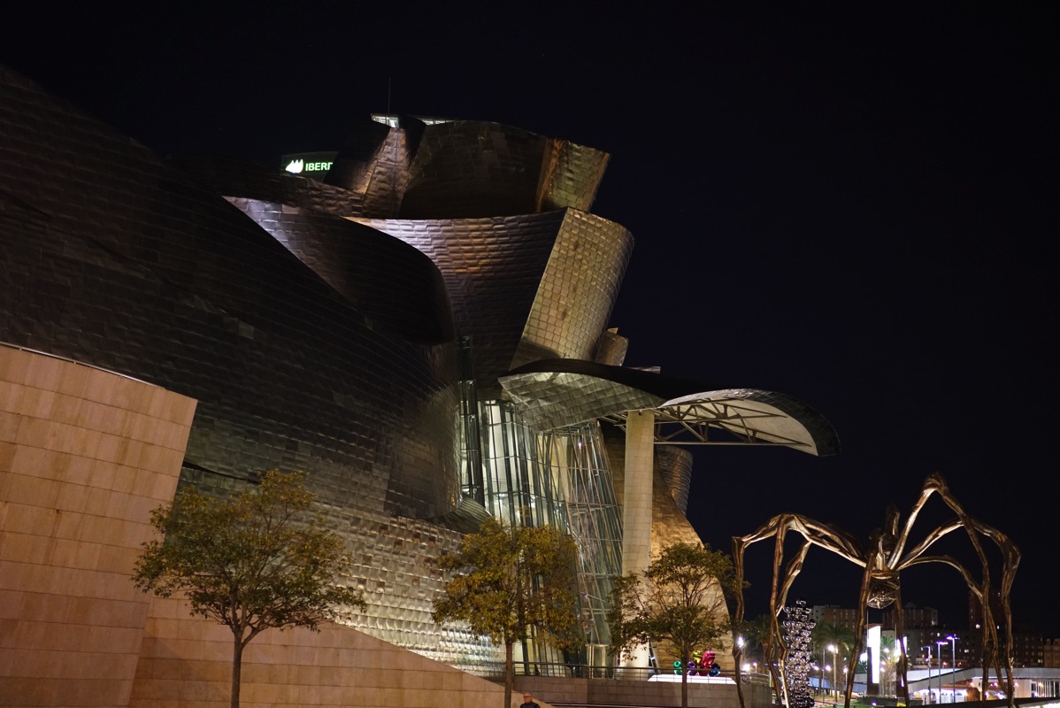 A nighttime photograph of the Guggenheim Museum in Bilbao, Spain, with the massive, bronze and steel spider sculpture 'Maman' visible in the foreground. The image captures a prominent artistic duo at the museum and the post's title, 'mum,' which is a play on the sculpture's name and its companion piece, 'Puppy,' from the same series.