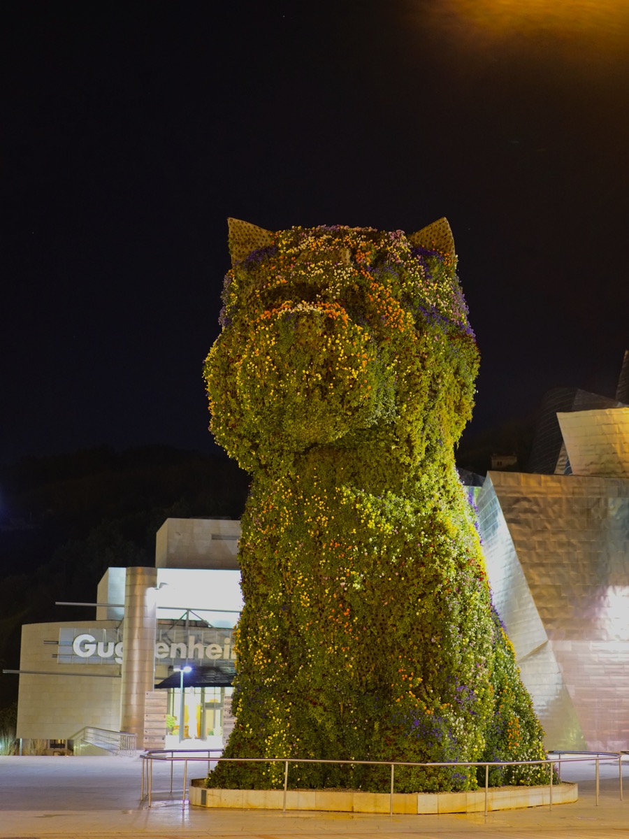 A vertical photograph of a massive dog-shaped sculpture, a living topiary made of green flowers and foliage, standing in front of the Guggenheim Museum in Bilbao at night. The sculpture, known as 'Puppy,' is an iconic landmark, and its presence adds a playful, whimsical touch to the post, which is titled 'pup'.