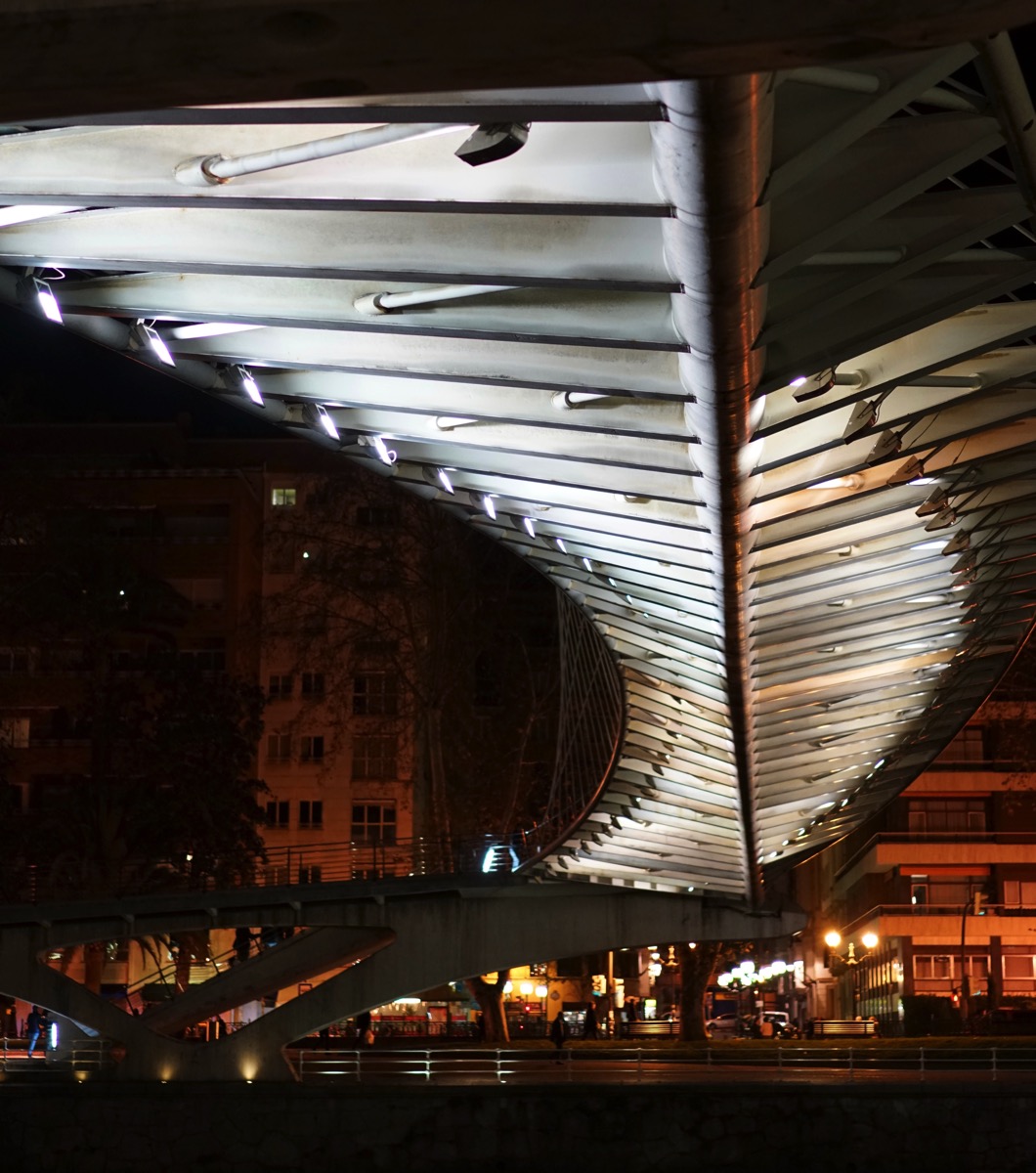 A dramatic, low-angle photograph capturing the curving underside of the Zubizuri Bridge in Bilbao, Spain, at night. The bridge's white, structural ribs are illuminated by a series of lights, creating a captivating pattern of light and shadow against the dark backdrop of the river and city lights. The image's perspective directly reflects the post's title 'under'.