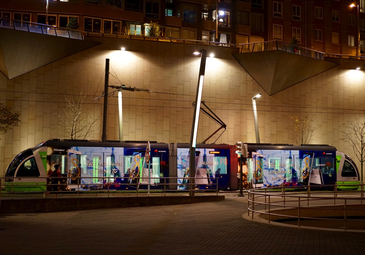 A vibrant night photograph of a modern tram in Bilbao, Spain, moving along its tracks. The brightly lit tram and its surroundings, including the city buildings in the background, reflect off the wet pavement. The image's composition and dynamic lighting capture the feeling of a late-night 'ride' in the city.