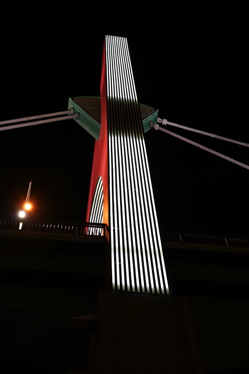 A dramatic, low-angle photograph of the abstract, fin-like architectural support of the Zubizuri Bridge in Bilbao, Spain, at night. The bridge's vertical supports are illuminated with bright, parallel lines of light, creating a strong contrast against the dark sky and reflecting the post's theme of an 'after dark' series.