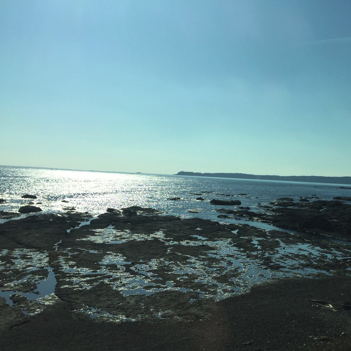 A wide-angle landscape photograph of the rugged, rocky coastline of the Kii Peninsula in Japan, taken on the Kumano Kodo pilgrimage route. The sun is shining brightly, casting a brilliant reflection on the surface of the hazy ocean water. The rocky shore is exposed by the low tide, and the sky is a clear, cloudless blue.
