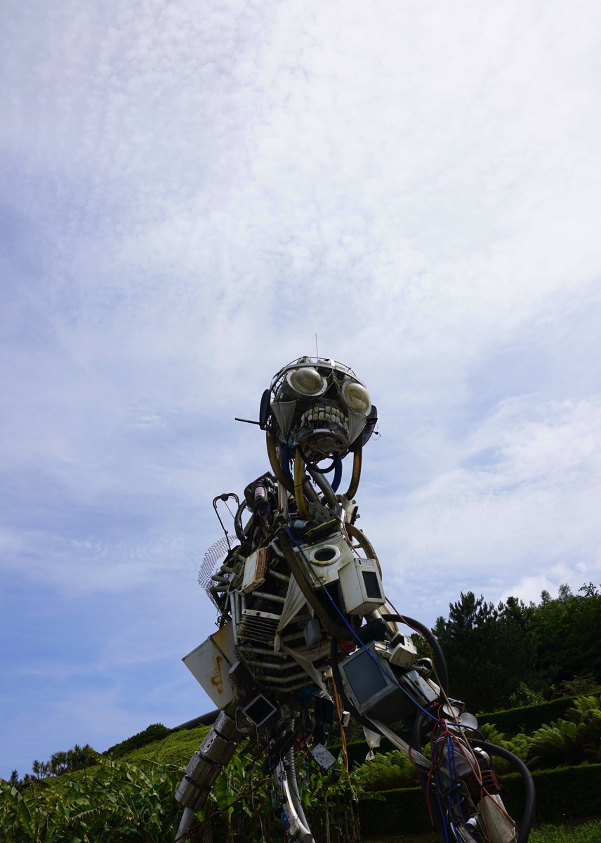 A vertical photograph of 'The WEEE Man,' a large sculpture of a humanoid figure made from metal and waste electronic materials, created by artist Paul Wager. The sculpture stands outdoors at the Eden Project in the UK.