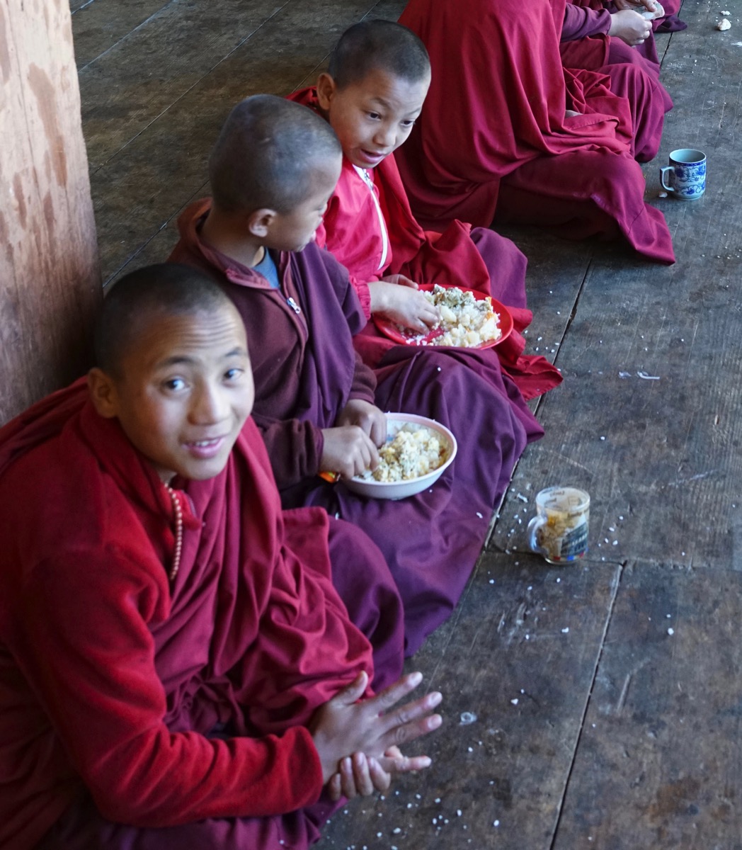 A photograph of three young Buddhist novices in red robes, sitting on a wooden floor and eating from metal bowls inside Gangtey Gonpa monastery in Bhutan. 