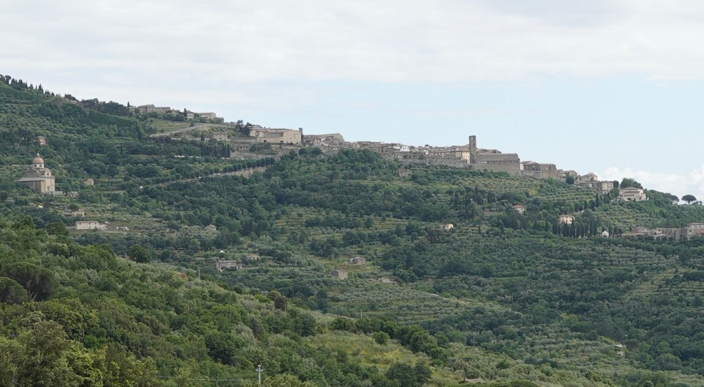 A mid-distance photograph of the ancient Tuscan hilltop town of Cortona, with its historical buildings and bell tower nestled into a lush green hillside dotted with cypress and olive trees. The image's quiet beauty and timeless feel serve as a visual counterpoint to the poem's comparison of Cortona's fame to the historical obscurity of Asciano.