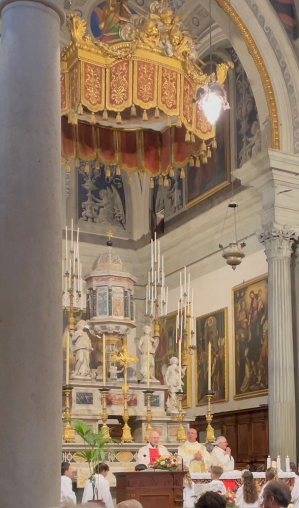A mid-distance photograph of the interior of the Cortona Duomo, focusing on the ornate pulpit and a section of the nave. Figures in the pews suggest a ceremony in progress, adding a human element to the grand architecture of the Tuscan cathedral.