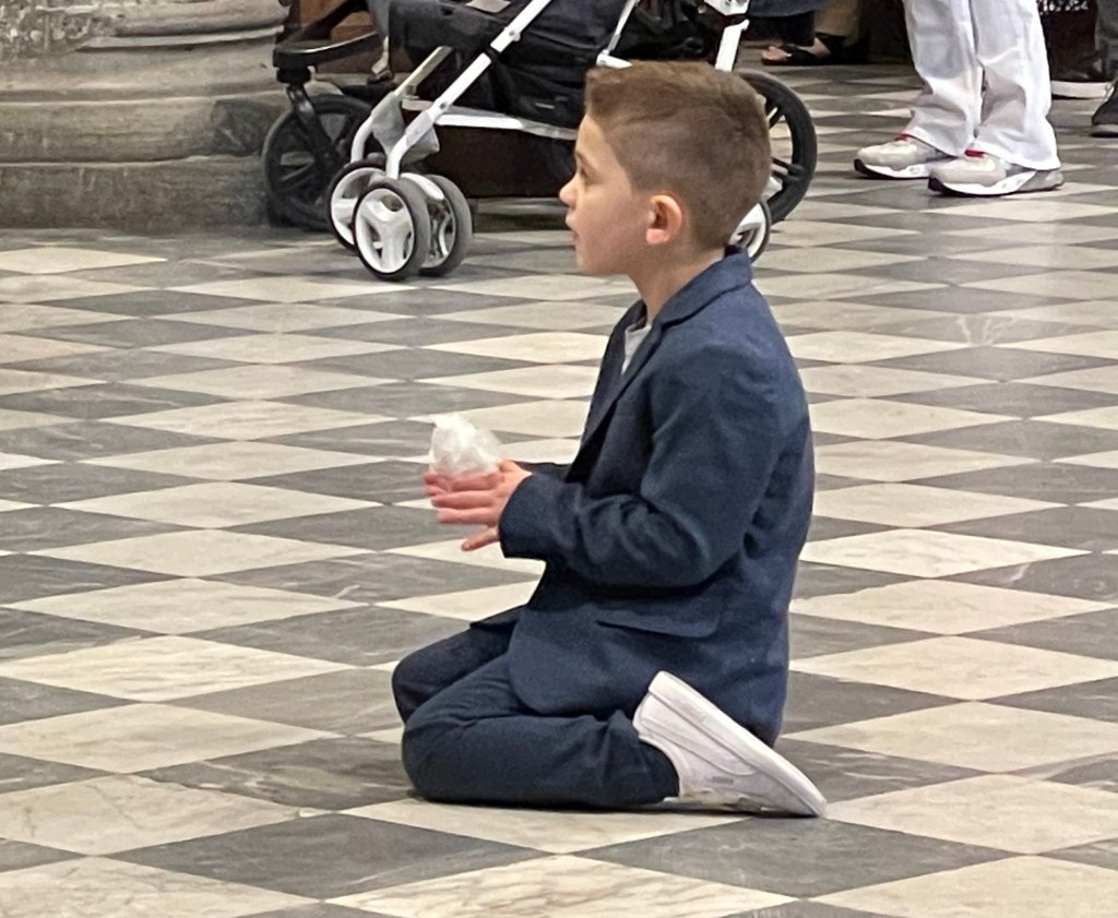 A close-up photograph of a young boy in a suit kneeling on the checkered floor of the Cortona Duomo. The image's solemn focus on the child reflects the poem's theme of a 'ceremony of first communion' and the personal significance of the experience.