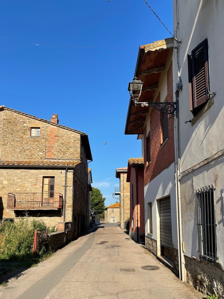 A photograph of a narrow, quiet cobblestone street in the small, medieval village on Isola Maggiore, an island on Italy's Lake Trasimeno. The image, with its ancient stone buildings and faded plaster walls, evokes a sense of history and the passage of time.